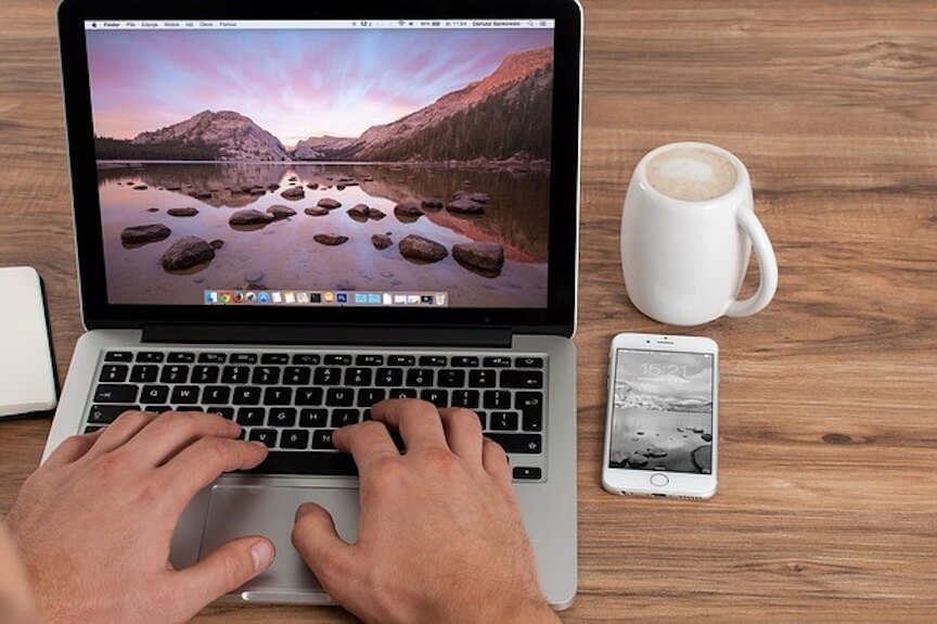 Person typing on a laptop with a smartphone and coffee on a wooden desk, representing remote work and digital productivity.