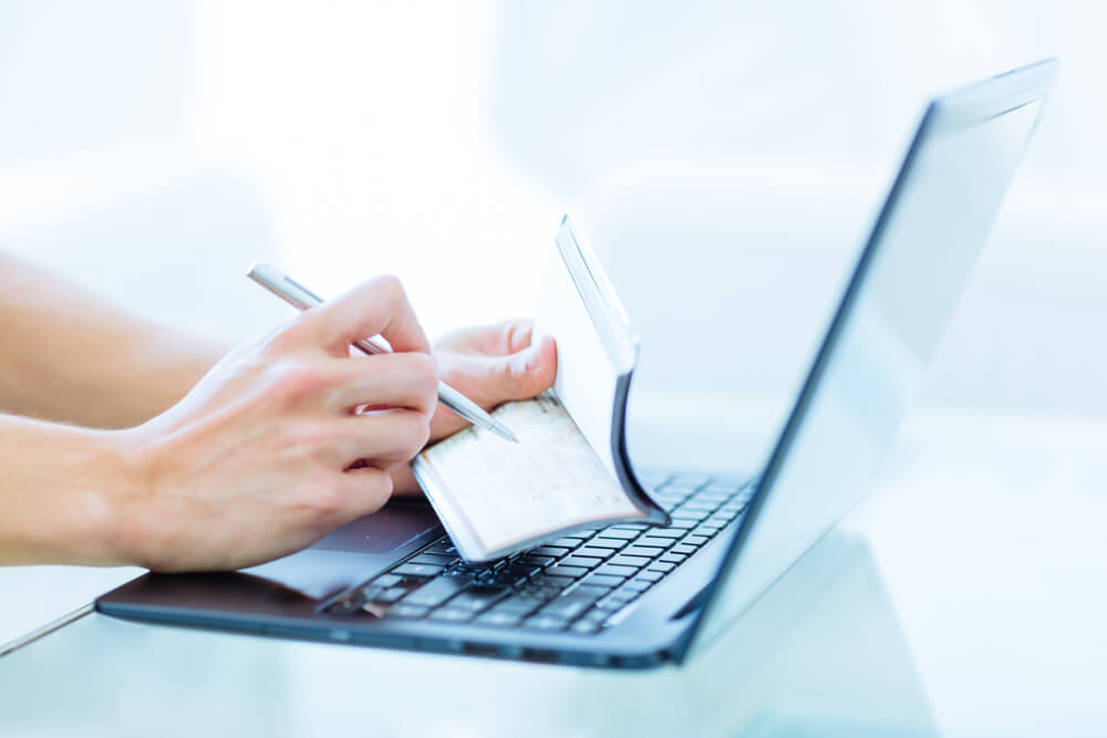 Closeup Of a Hands Writing a Personal Bank Cheque Using a Pen While Working on a Laptop Computer