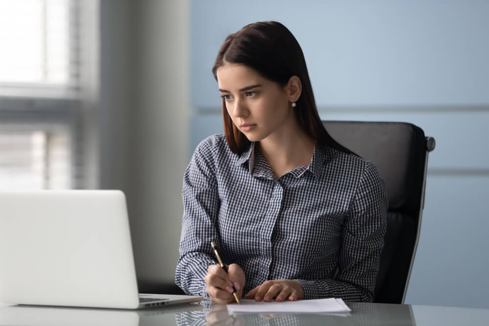 Concentrated Young Businesswoman Writingon Paper Watching Business Webinar on Laptop