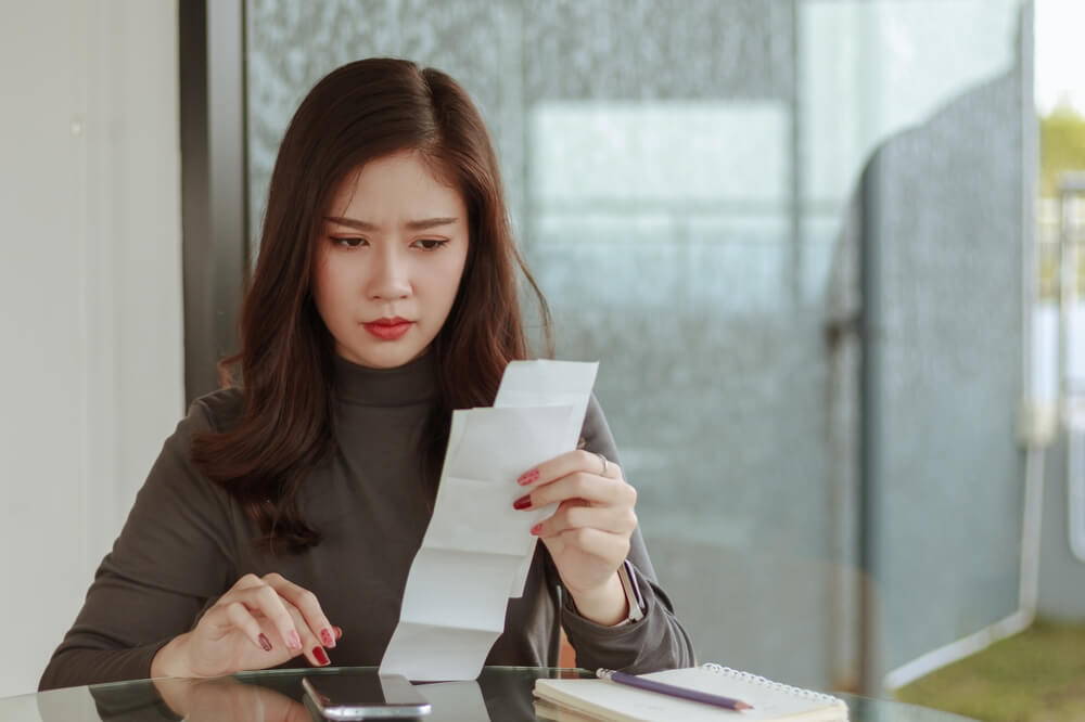 Young Woman Look At Pay Bill Holding Ache At Work. Tired Woman Employee Looking At Report On Paper Having Headache.